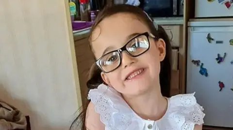 Lancashire Police Six-year-old Millie Gribble with long dark brown in a pony tail wearing a frilly white blouse and black glasses standing in a kitchen. Her head is tipped to the side and she has a big smile. 
