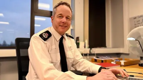 Zara Healy/BBC Chief Constable Paul Gibson seated at a desk. He is wearing police uniform and smiling at the camera, There is a large window in the background.