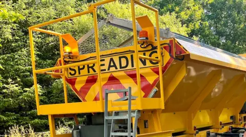 A close-up of the back of a yellow gritting truck parked on the side of a road. The back of the truck has "spreading" on the back, on top of a red and yellow pattern. Trees can be seen in the background, behind the truck.