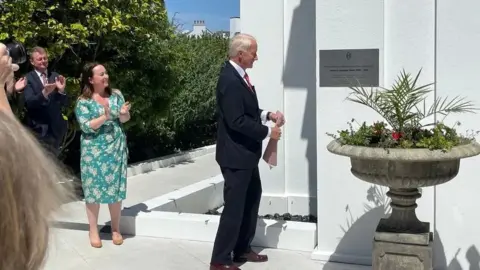 BBC A man with grey hair and a dark suit opens a white building surrounded by plants.