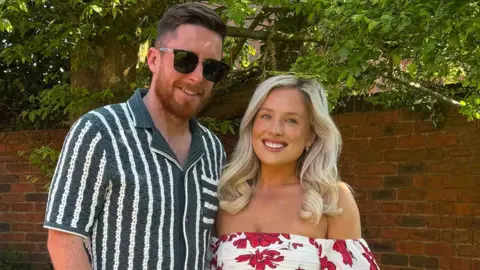 Two people, Kat and Adam standing smiling in a garden. She wears a summer flowery dress and has blonde hair. he has brown hair and a beard and wears white trousers, a stripy shirt and sunglasses.