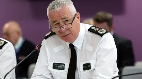 PA Media Police Service of Northern Ireland Chief Constable Jon Boutcher is speaking into a microphone. He is sitting in front of a purple wall with three men behind him, who are out of focus. He is wearing a white shirt which has police badges on both shoulders and also a black tie.