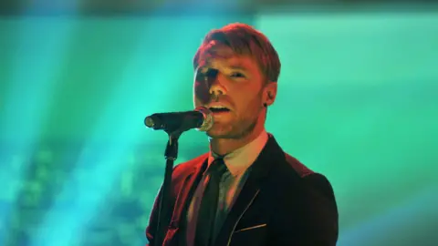 Ronan is singing into a microphone as blue lights shine around him. He is wearing a suit jacket, tie and white shirt. He has short hair and some facial hair.