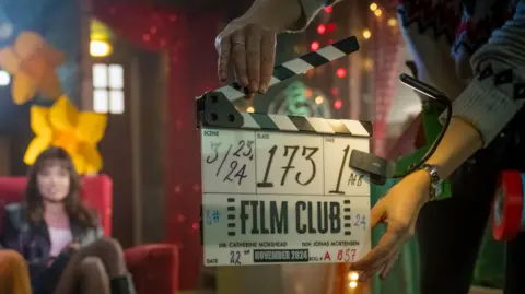 BBC/Gaumont/Ben Blackall An unidentified worker holds a TV clapper board with details for a shoot for Film Club. It is in front of a scene featuring the actress Aimee Lou Wood sat on a sofa in a colourfully-lit room with huge daffodil emblems on the background.