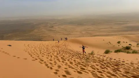 Adam Van de Velde A wide expanse of orange coloured sand with a man wearing a cap running with his arms open wide and five other runners in the distance.