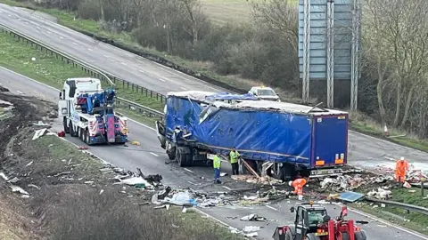 Accident scene showing a blue lorry in the central reservation surrounded by debris and emergency workers cleaning up with a vehicle recovery truck parked on the road 