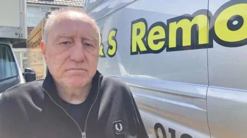 Mousumi Bakshi/BBC A man is looking concerned while standing next to a silver van with a yellow logo on it which says "Remo".