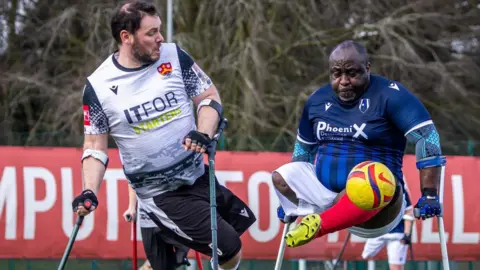 Footballers are on a pitch. Both have crutches and one has his leg in the air as he kicks a ball.