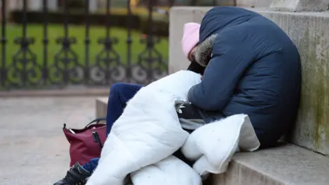 Two people on a concrete step hunched over. One is wearing a blue hooded coat, with a fur trim on the hood and a pair of black Adidas trousers, holding on to a white duvet. The other is wearing a baby pink hat, dark blue jeans and black walking boots. There is a burgandy bag beside their foot.