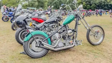 Hornsea Bike Show A green motorcycle with extended forks and long handle bars. There are other motorcycles in a row behind it and people in the distance.