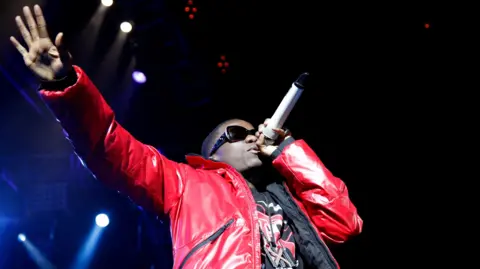 Tinchy Stryder performs on stage at night. He is wearing a red coat, a black and red t-shirt, and big sunglasses. He is holding a microphone to his mouth and has his other arm extended. Stage lighting can be seen behind him.