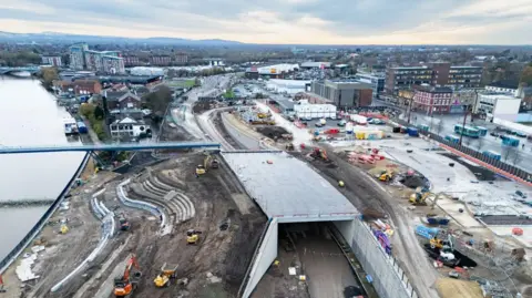 Construction work along the waterfront area of Stockton. A number of diggers and similar vehicles are on the site. To the left of the image, curved granite steps are being put in place to create a terraced area near where a footbridge spans the River Tees.
