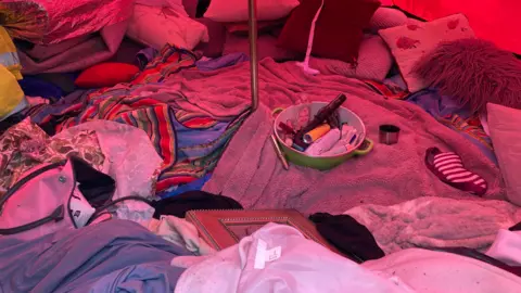 The insides of a tent, showing toiletries, old bottles of deodorant, clothes, cushions and towels.