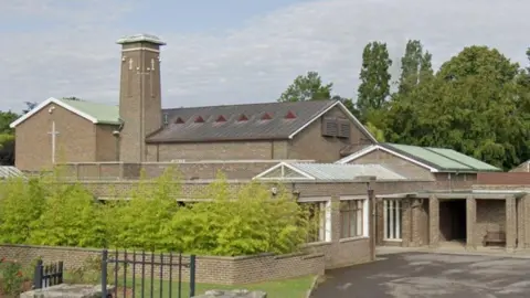 The picture show Porchester Crematorium, a building with Christian crosses imprinted onto the brickwork. Part of the building is a tall tower that rises above the rest of it. In front of the building is a lush green bush. 