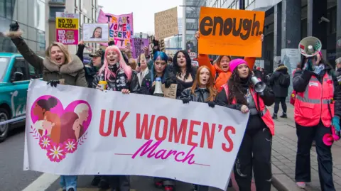A group of protesters walk down a London street. They hold up a large banner saying UK Women's March. They hold up handmade signs saying 'enough' and 'being a woman shouldn't be a death sentence'.