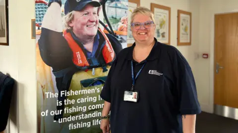 BBC Hayley Hamlett from the Fishermen's Mission smiles at the camera in front of a banner which has a smiling fisherman on in