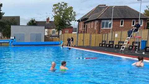 BBC People swimming in the blue water of an outdoor swimming pool with a lifeguard on a ladder to the right and red-brick buildings and trees in the background behind a wooden fence.