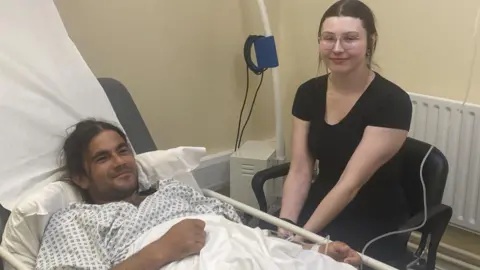 Paul Byrne A man in a hospital bed and gown, beside him a woman sits on a chair