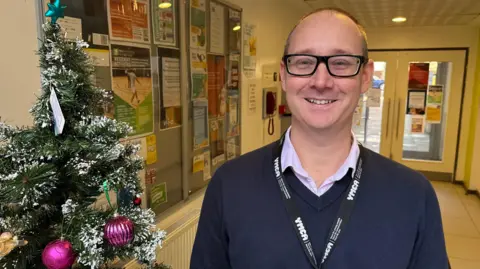 A man wearing a navy v neck jumper smiling at the camera. He is wearing glasses and standing next to a Christmas tree