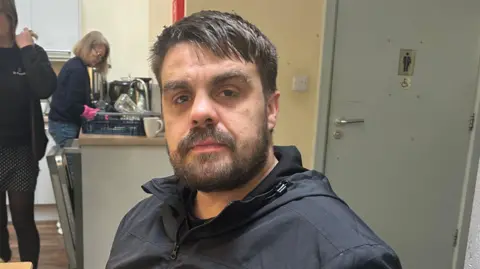 A man with dark brown hair and a beard wearing a black jacket