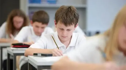 Four pupils are sitting in a row behind each other, they are wearing white school uniform shirts and are writing