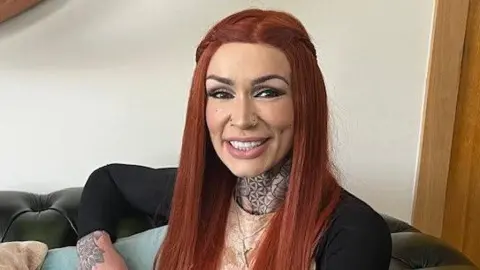 Michelle Moffatt has long ginger hair and smiles at the camera. She has neck and hand tattoos and appears to be sitting on a couch in a living room.