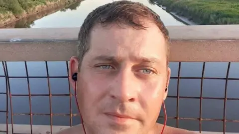 Handout James Miller is pictured in a selfie. He is on a bridge or walk way across a river. He is wearing headphones. He has blue eyes and short brown hair.