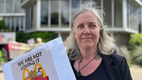 Emma Russell wearing a black blazer and holding a sign saying 'we are not loving it'