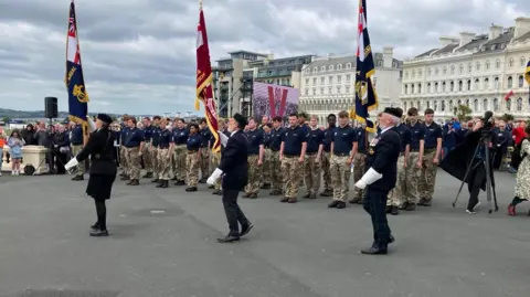 A group of military people stood in formation with flags being carried on Plymouth Hoe.