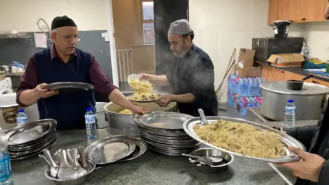 Two men are cooking and a third man stands to the side holding a large plate of food. One of them is piling the food on to a plate. There are dozens of large silver plates on the table.