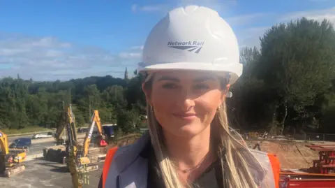 Olivia Boland stands on a grass bank above the site where preparatory work for the bridge replacement is under way