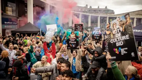 Getty Images A large crowd of people huddled together outside holding signs and flags. Red and blue smoke billows in the background