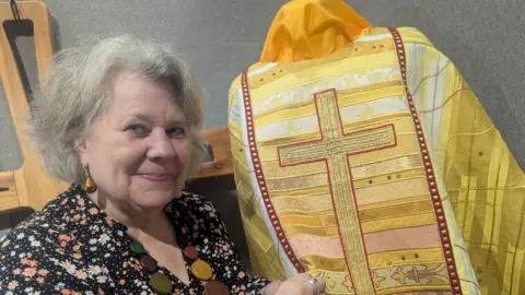 Christian Resources Exhibition Juliet Hemingway with grey hair wearing a flower-patterned dress and holding a yellow clerical cloak with a large cross embroidered onto it.