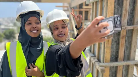 Saadia Ahmed Omar (right) takes a photo of herself and Fathi Mohamed Abdi (left) atop a building under construction in Mogadishu. They are both wearing hard hats over their headscarves and are in high vis yellow vests. Ms Omar makes the victory sign as she takes the photo.