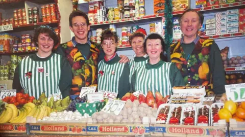 Supplied A group of staff lined up in matching uniform behind boxes of fruit and veg. Female workers are wearing outfits with vertical green and white stripes. The two men in the shot are wearing waistcoats covered in various fruits. 