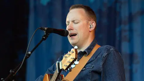 A man standing onstage with an acoustic guitar, singing into a microphone with his eyes closed. He has cropped dark hair and is wearing denim.