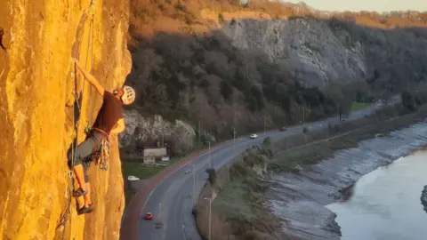 Lucas Harcombe A climber hangs by one arm on a cliff. A road runs beneath it and a river is visible on the right. The Clifton Suspension Bridge is visible in the background.