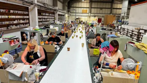A long white table stretching down the middle, with colourful mugs on it. On either side of the table are women sitting at desks and painting mugs. There are oak shelves of mugs lining the walls.