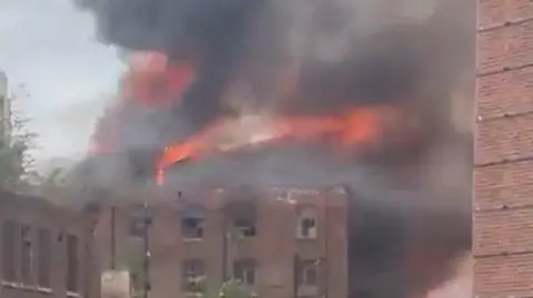 X Image shows flames and thick smoke rising from the roof and upper floors of the Hotspur Press building in Manchester, a Victorian-era red-brick warehouse.