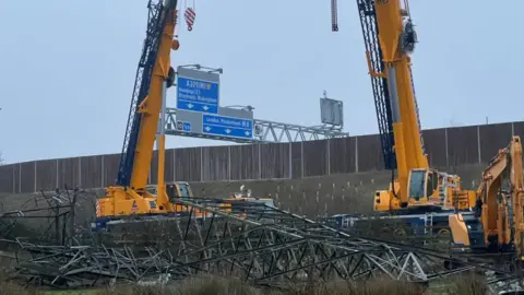 A general view of two large cranes over a pylon that has been brought down. The M4 sign is visible behind it on a grey day.
