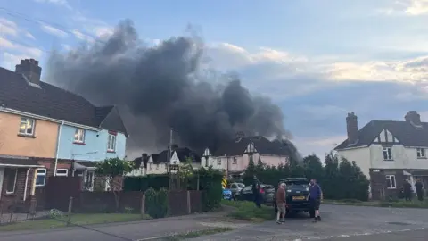 A large plume of black smoke coming from a house on Jubilee Road in Lydney. The photo has been taken on a cloudy evening. There are some people standing by some cars on the street.