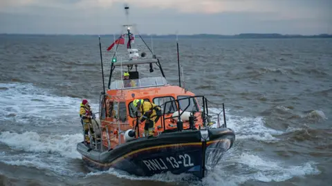 Bridlington RNLI/Mike Milner A lifeboat with three crew members in view, with one of them at the helm. "RNLI" is written on the side and the lifeboat is heading through water