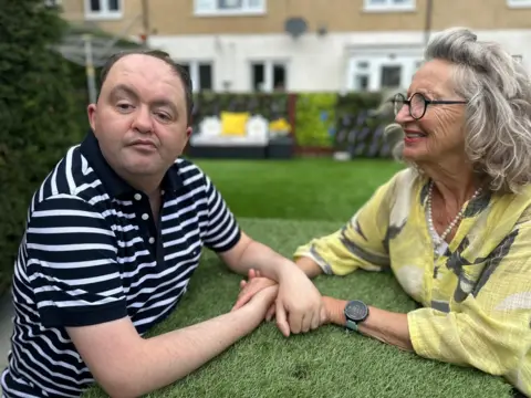 BBC Keith Fisher, who has cerebral palsy, sits at a garden table with his mum Kim. Keith wears a navy and white striped t-shirt and his mum is in a bright yellow shirt, with a pearl necklace and black circular-rimmed glasses.