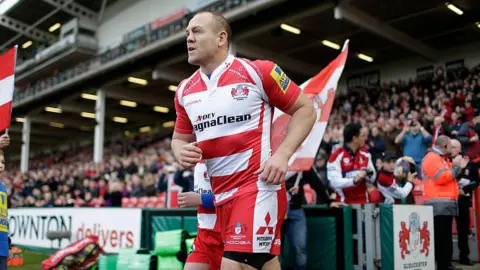 Getty Images Mike Tindall, a Gloucester Rugby player wearing the team's cherry and white kit, runs out onto a pitch beside a younger mascot. There are fans in the background in the stands, cheering and clapping him on.