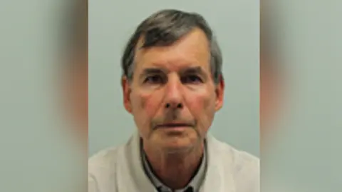 A mugshot of a man with brown hair, a white jacket and grey shirt.