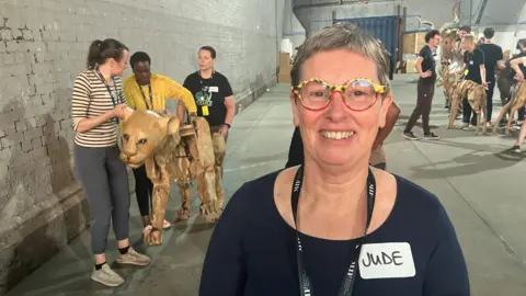 Photograph of Jude Battersby in front of some of the animal puppets underneath a series of railway arches in Manchester city centre. 