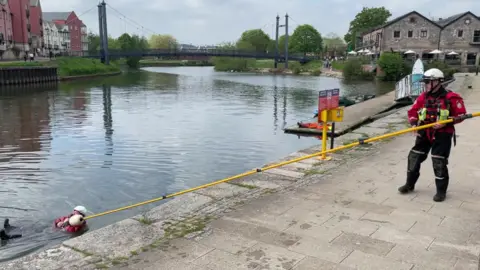 A person in red and black outfit and white hat holds a long yellow pole from a quayside to a person holding the pole in a river.