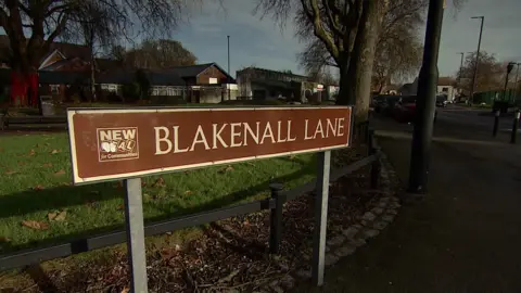 A brown road sign with white lettering on Blakenall Lane. The sign also feature the 'new deal for communities' logo.