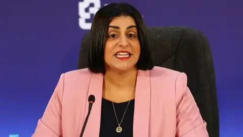 PA Media Shabana Mahmood sits in a high backed grey chair and speaks into a mic in front of a blue background