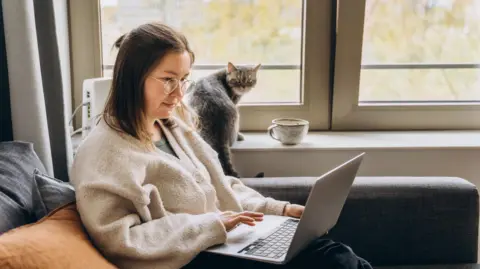 Getty Images Woman sitting on a sofa using a laptop. A cat and a large ceramic mug are on the windowsill behind her. She has long brown hair, and is wearing glasses and a grey cardigan.